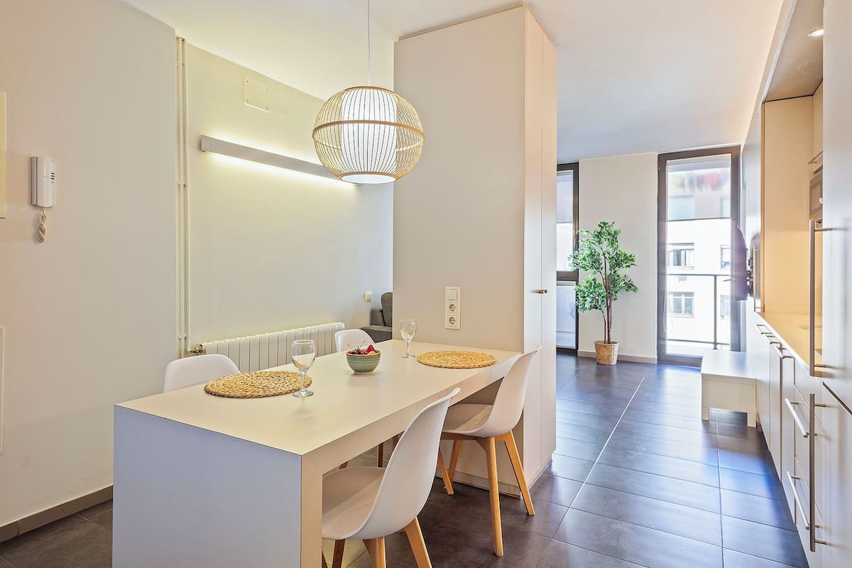 A modern kitchen with a white countertop, two white chairs, a hanging light fixture, and a potted plant.