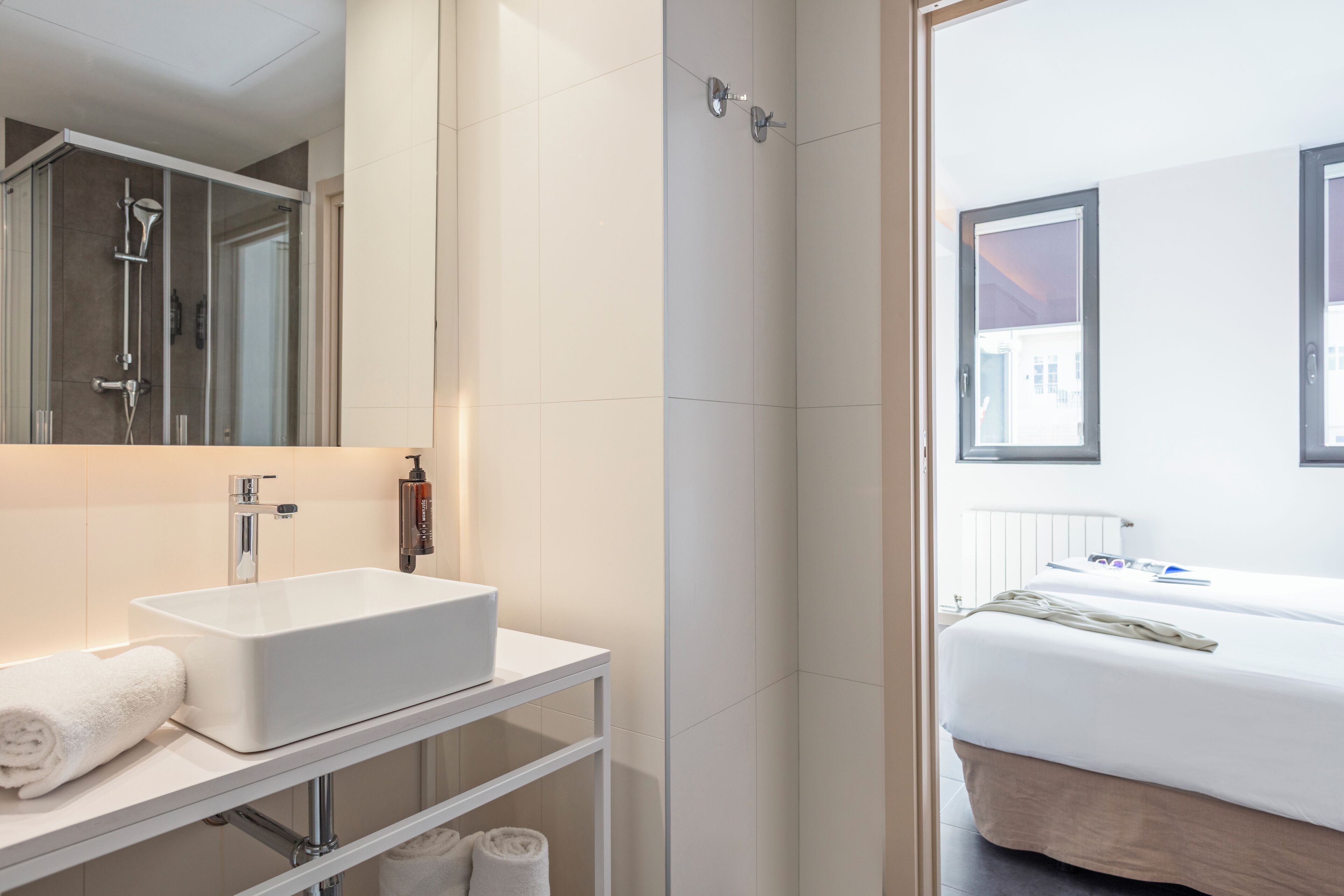Une salle de bain moderne avec un lavabo blanc, un miroir et une douche. On y trouve un lit avec un matelas blanc et une fenêtre donnant sur des immeubles.