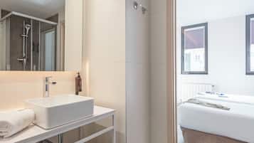 Une salle de bain moderne avec un lavabo blanc, un miroir et une douche. On y trouve un lit avec un matelas blanc et une fenêtre donnant sur des immeubles.