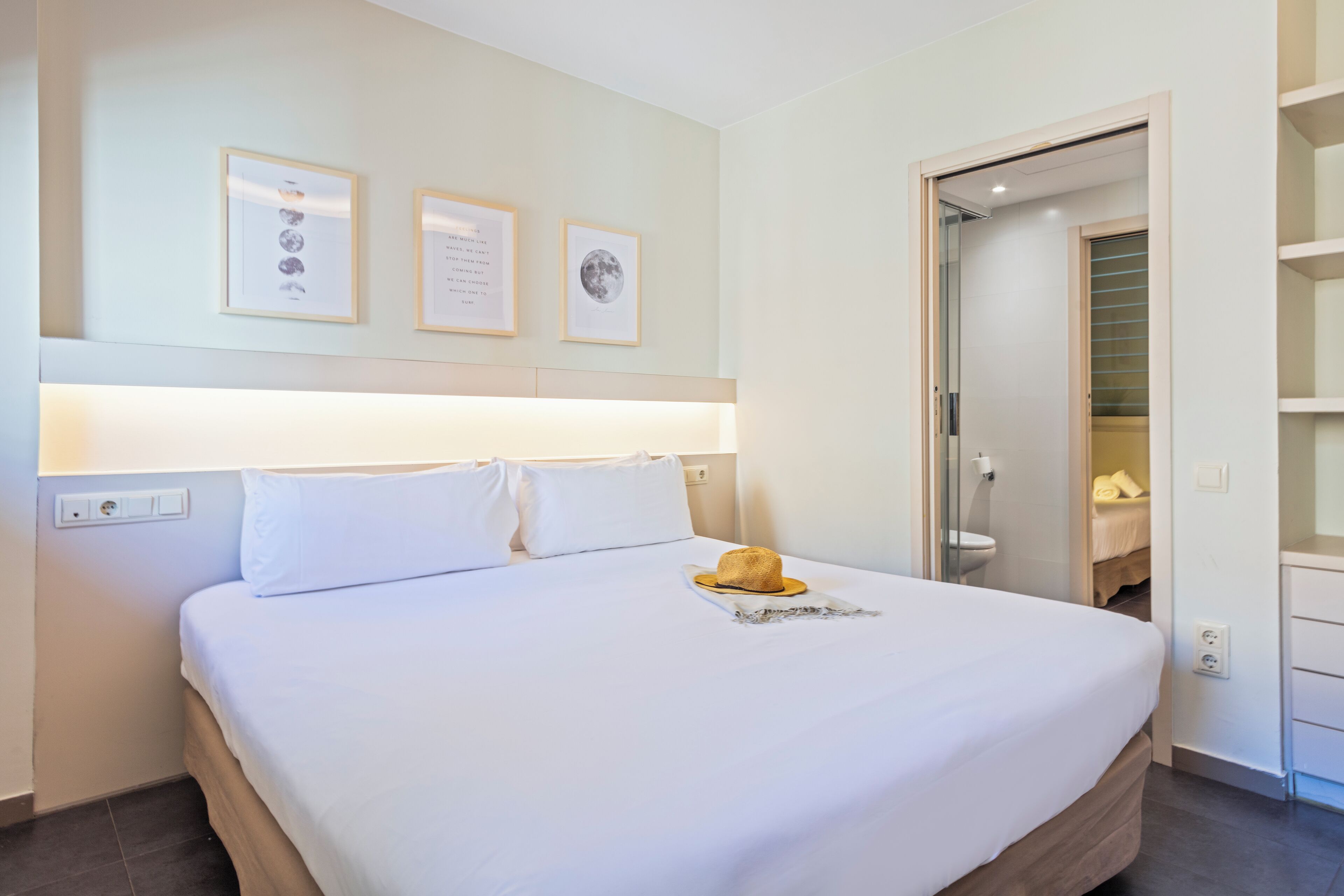 Una cama bien hecha con sábanas blancas, cabecera de madera y un sombrero sobre la almohada. En la pared, encima de la cama, hay cuadros enmarcados.