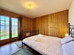 Une chambre avec un mur lambrissé en bois, un lit recouvert de draps blancs, une vue à travers une grande fenêtre et un luminaire au plafond.