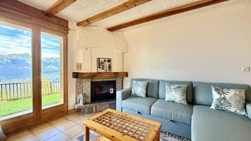 Una sala de estar con chimenea, un sofá, una mesa de centro de madera y vistas a montañas a través de amplios ventanales.