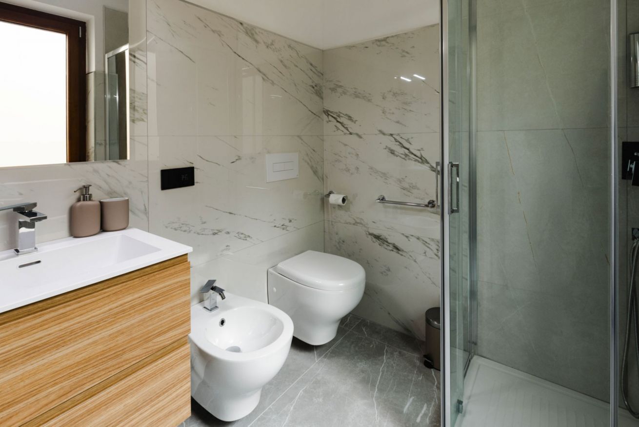 A modern bathroom with a marble wall, a wooden vanity, a bidet, a toilet, and a glass shower enclosure.