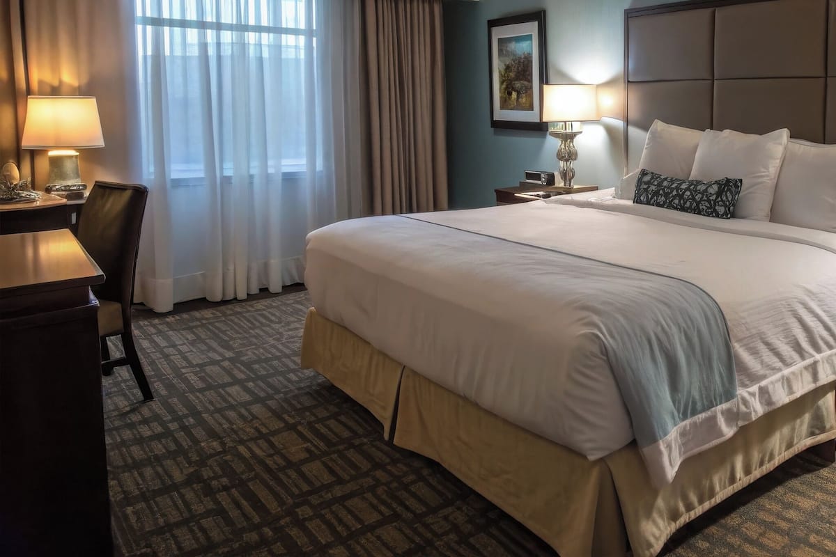 A hotel room with a large bed, a desk with a lamp, a chair, and a window with curtains.