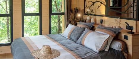 A wooden cabin bedroom with a bed, large windows, and a tree branch artwork on the wall.
