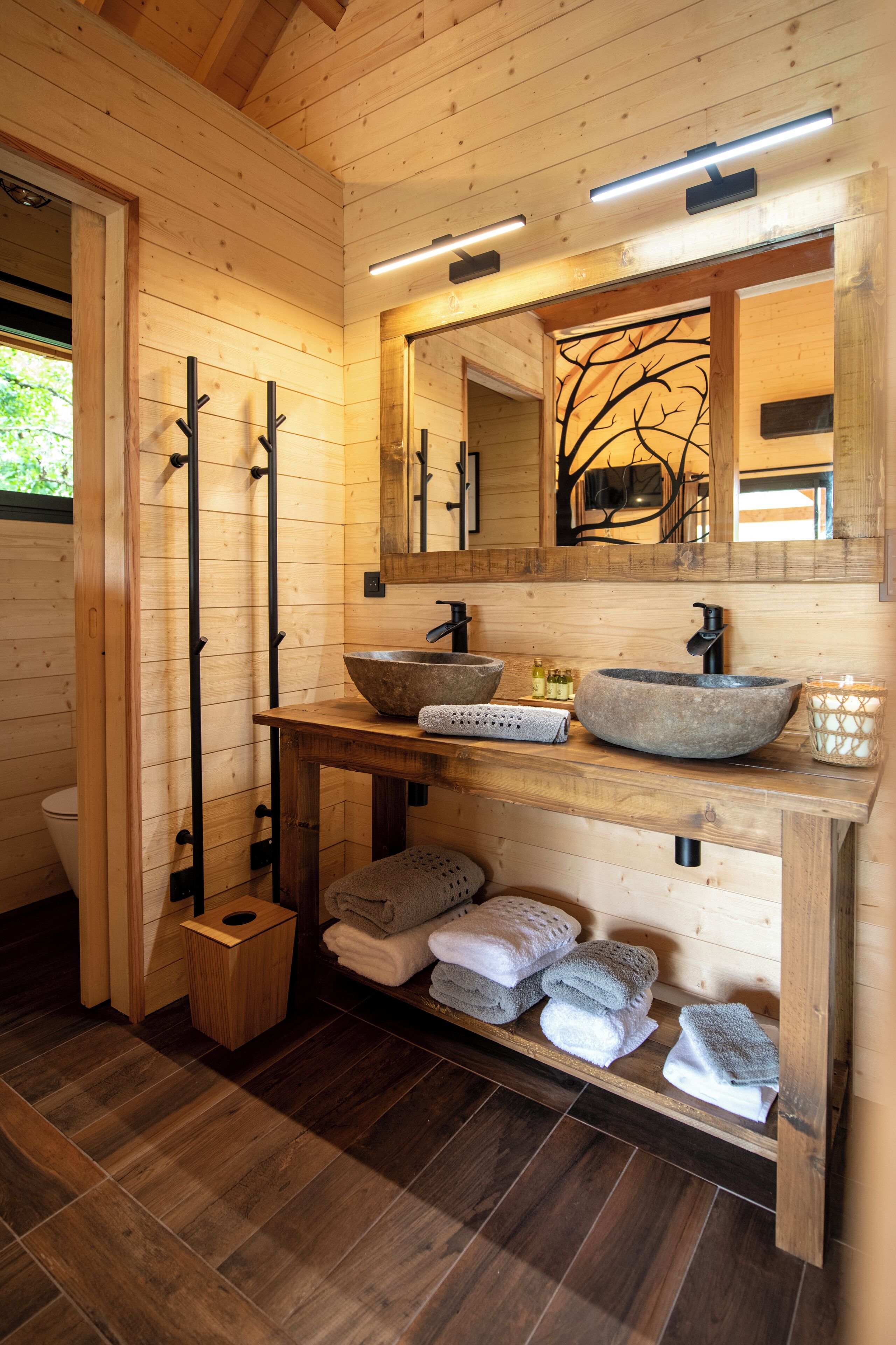 A wooden cabin bathroom with a wooden vanity, two sinks, a mirror, and a shelf with towels.
