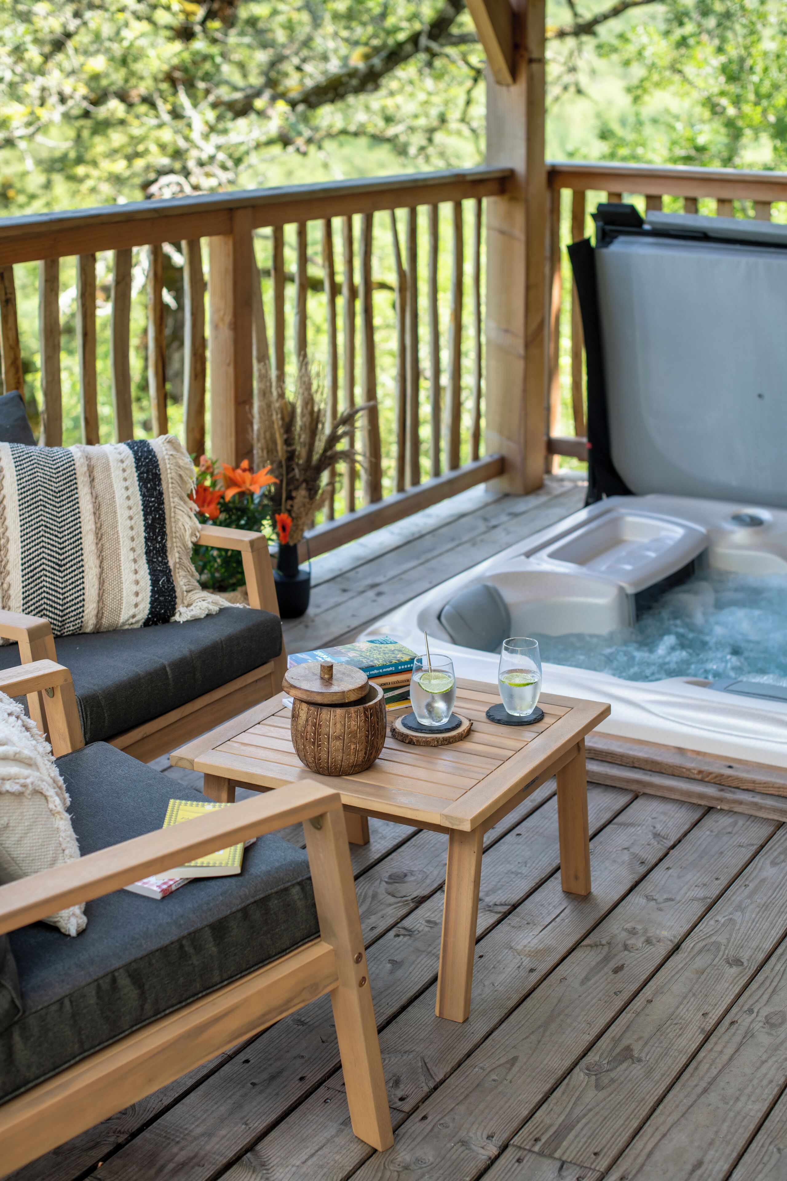 Une terrasse en bois avec un canapé, un jacuzzi et une table avec des verres et un livre.