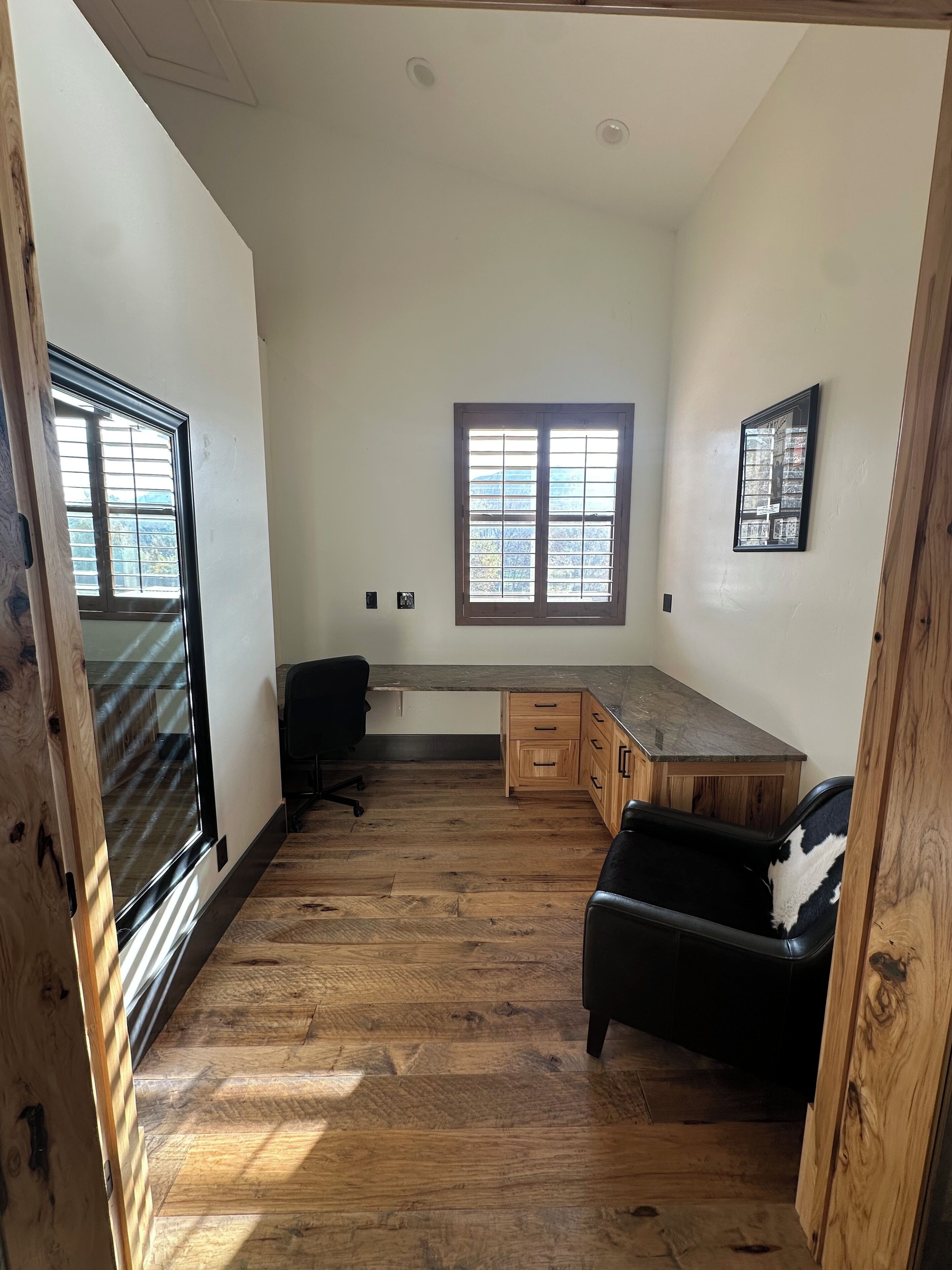 A spacious room with a large window, wooden flooring, a desk, a chair, and a couch.