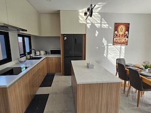 Eine moderne Küche mit Holzschränken, einer eingebauten Mikrowelle und einem Kühlschrank. Es gibt eine Essecke mit einem Tisch für zwei Personen, Stühlen und einem dekorativen Buddha-Gemälde.