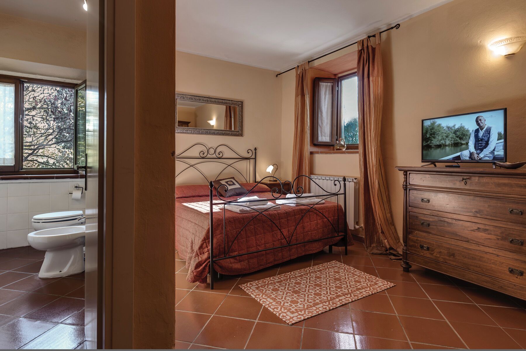 Une chambre à coucher comprenant un lit, une commode, un téléviseur, une fenêtre avec des rideaux, et une salle de bain avec des toilettes et une baignoire.