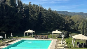 Ein Swimmingpool mit Liegestühlen und Sonnenschirmen, umgeben von viel Grün und Hügeln.
