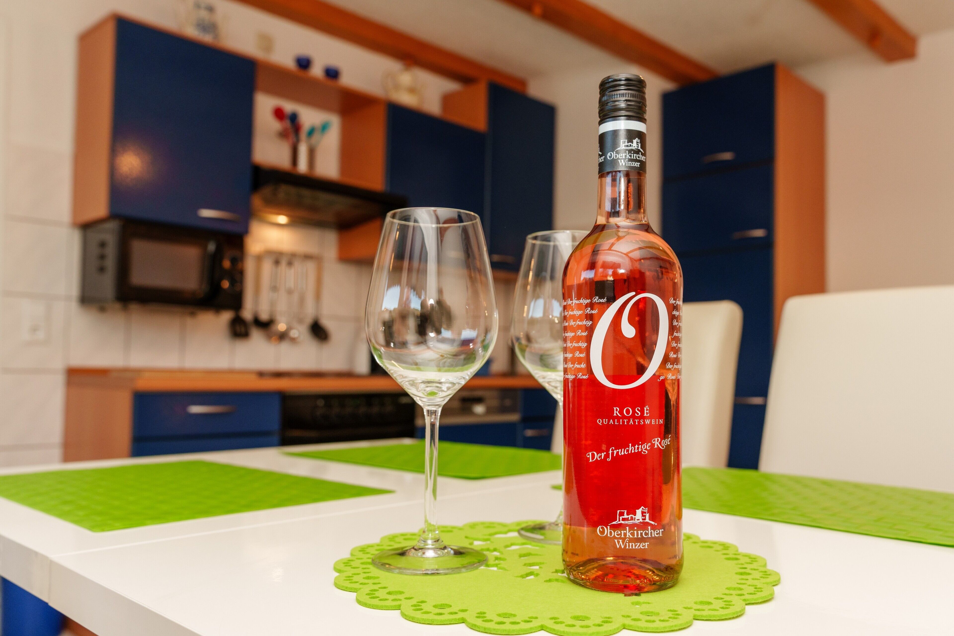 Una botella de vino rosado y dos copas sobre una mesa en una cocina moderna con armarios azules y un posavasos verde.