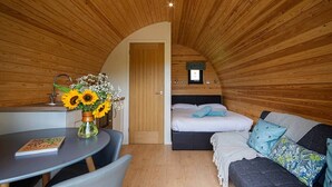 Una habitación acogedora con techo de madera, una cama con ropa de cama azul, un sofá con cojines estampados, una mesita con un libro y un jarrón con girasoles.