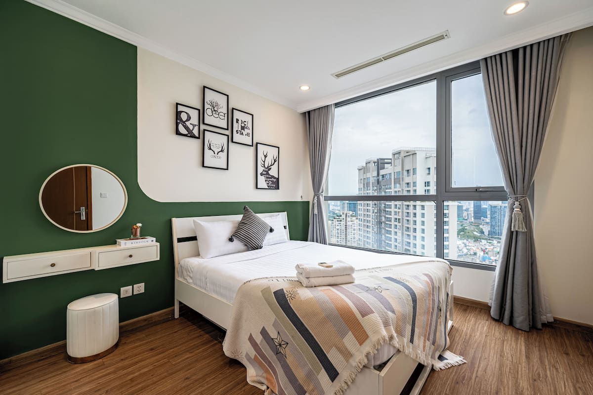 Ein modernes Hotelzimmer mit großem Fenster, einem Bett mit gestreifter Decke, einem runden Spiegel und Bildern an der Wand.