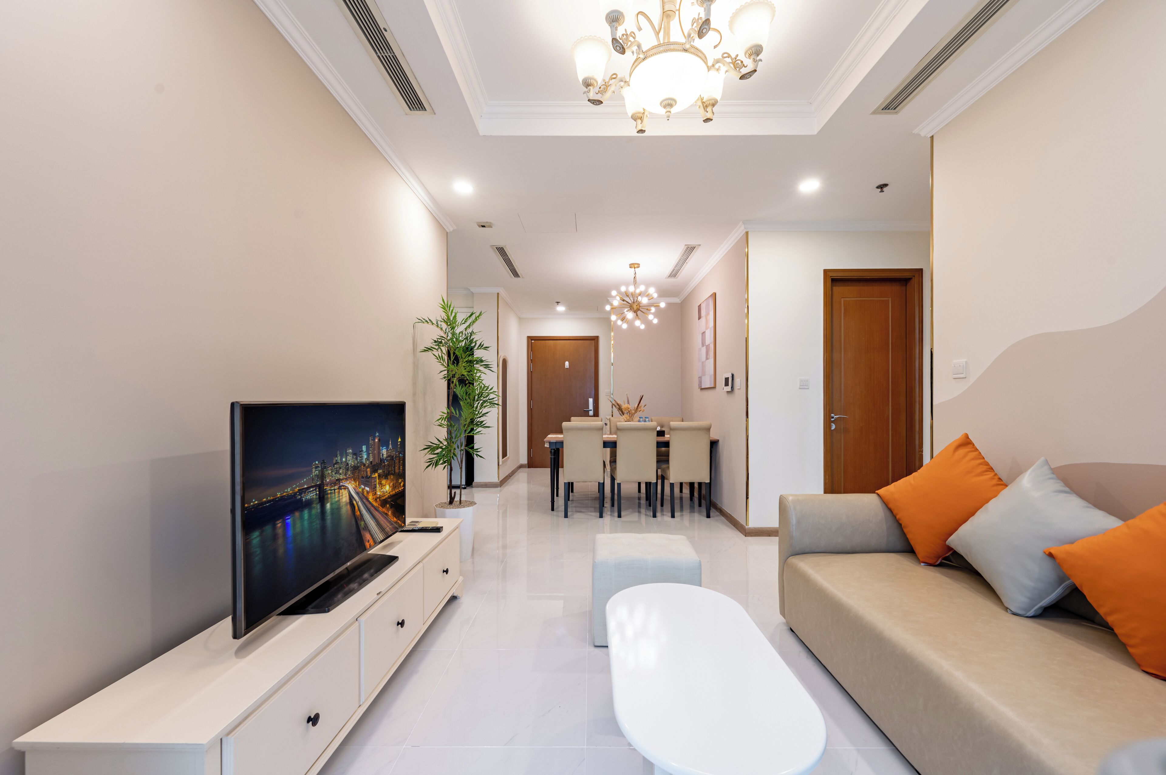 A modern living room with a flat-screen TV, a sofa with orange cushions, a dining area, and a chandelier.