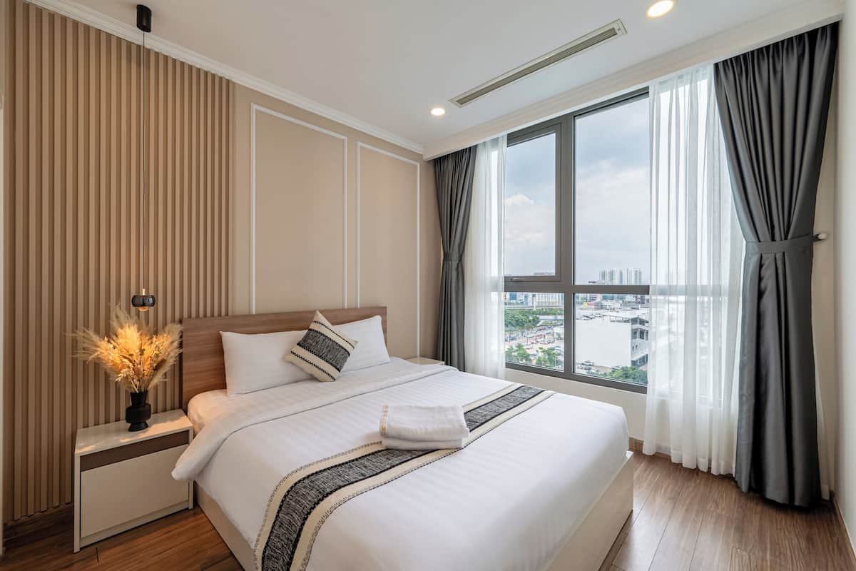 Ein Hotelzimmer mit einem großen Bett, einem Nachttisch mit Lampe und Blick auf die Stadt durch ein Fenster. Außerhalb des Fensters ist ein Stadtbild zu sehen.