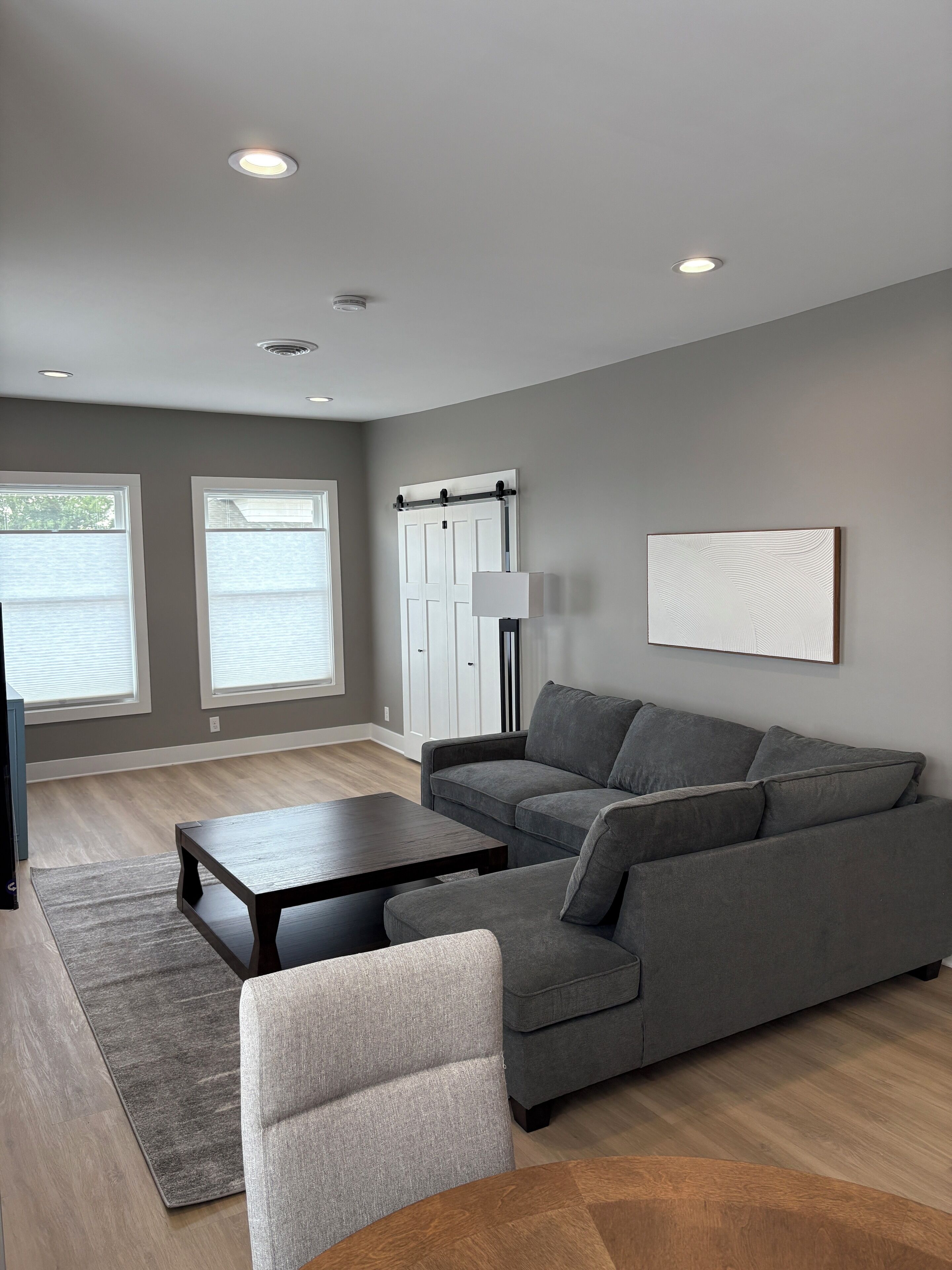 A modern living room with a grey sofa, a wooden coffee table, and a grey armchair. There are two windows with blinds, a white door, and a floor lamp.