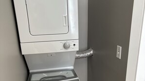 Una lavadora y secadora blancas y compactas ubicadas en un rincón de una habitación con piso de madera y paredes grises.