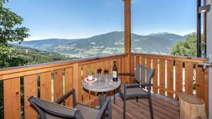 Een houten terras met een tafel, stoelen, een fles wijn en uitzicht op bergen.