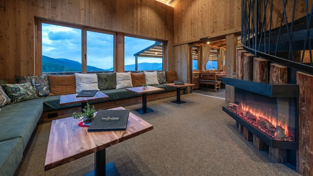 Ein Holzinnenraum mit einer Couch, einem Tisch, einem Kamin und Blick auf die Berge.
