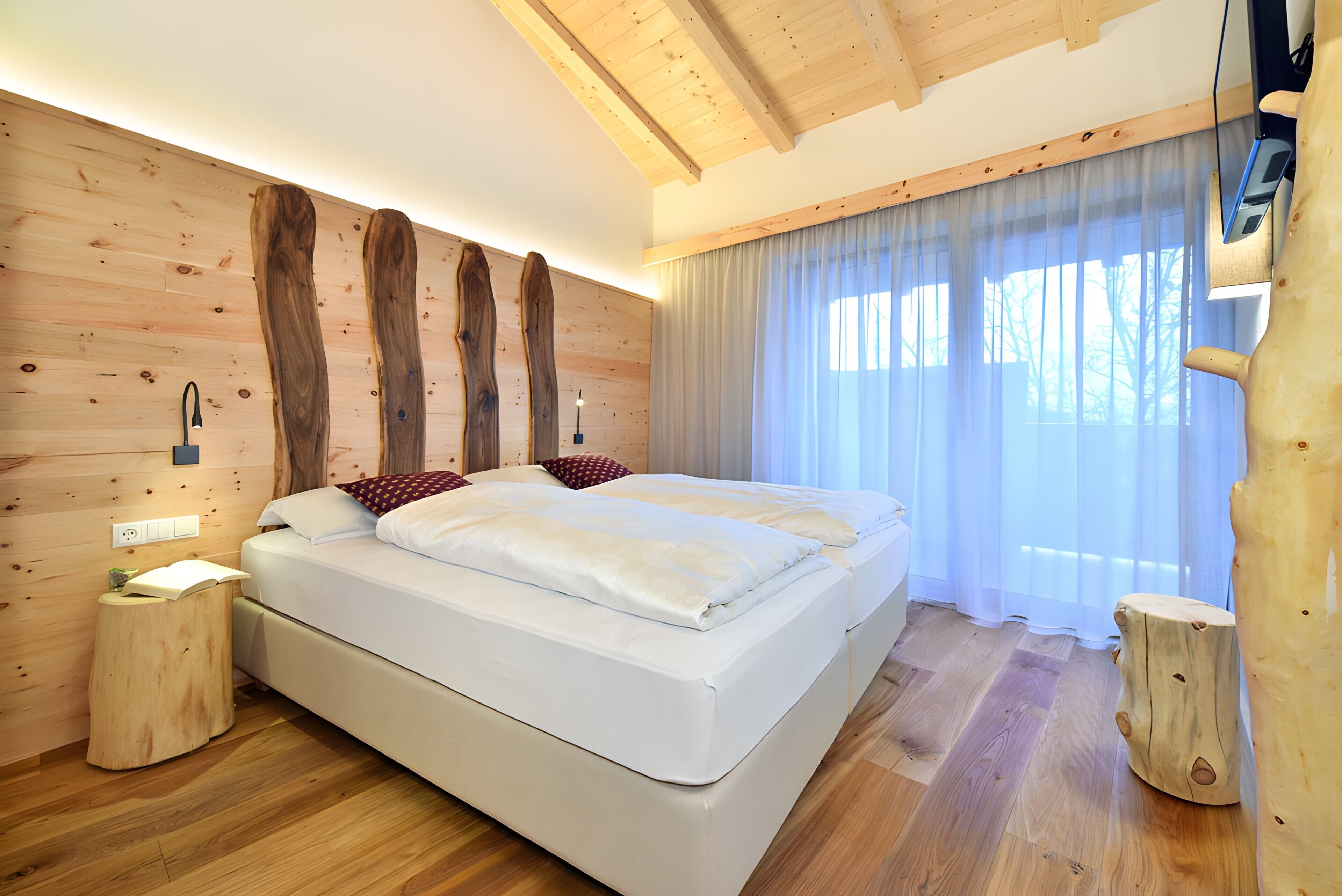 A modern bedroom with a large bed, wooden wall paneling, a bedside table, and a window with sheer curtains.