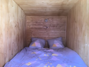 Um quarto pequeno de madeira com uma cama de solteiro, duas almofadas e um interruptor de luz visível na parede.