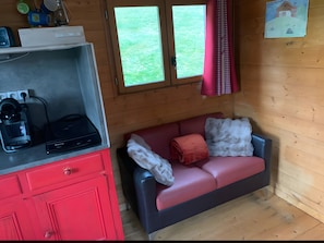 Quarto compacto com cozinha compacta, sofá vermelho com manta branca e janela com vista para vegetação.