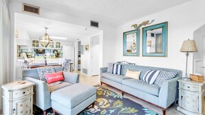 A living room with a sofa, ottoman, and side tables, a patterned rug, and a kitchen in the background. - 🏖️BeachFront-2 Pools-Great Location-Pinnacle Port PH36🏖️ (Panama City Beach)