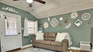 A living room with a brown sofa, a patterned rug, decorative wall art, and a ceiling fan. - Hidden Oaks | The Cottage | Unit 1 (Birch Run Township)