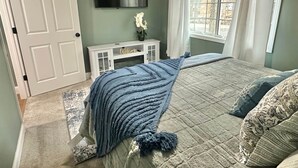 A bedroom with a bed, a TV, a console table, and a window with curtains. - Hidden Oaks | The Cottage | Unit 1 (Birch Run Township)