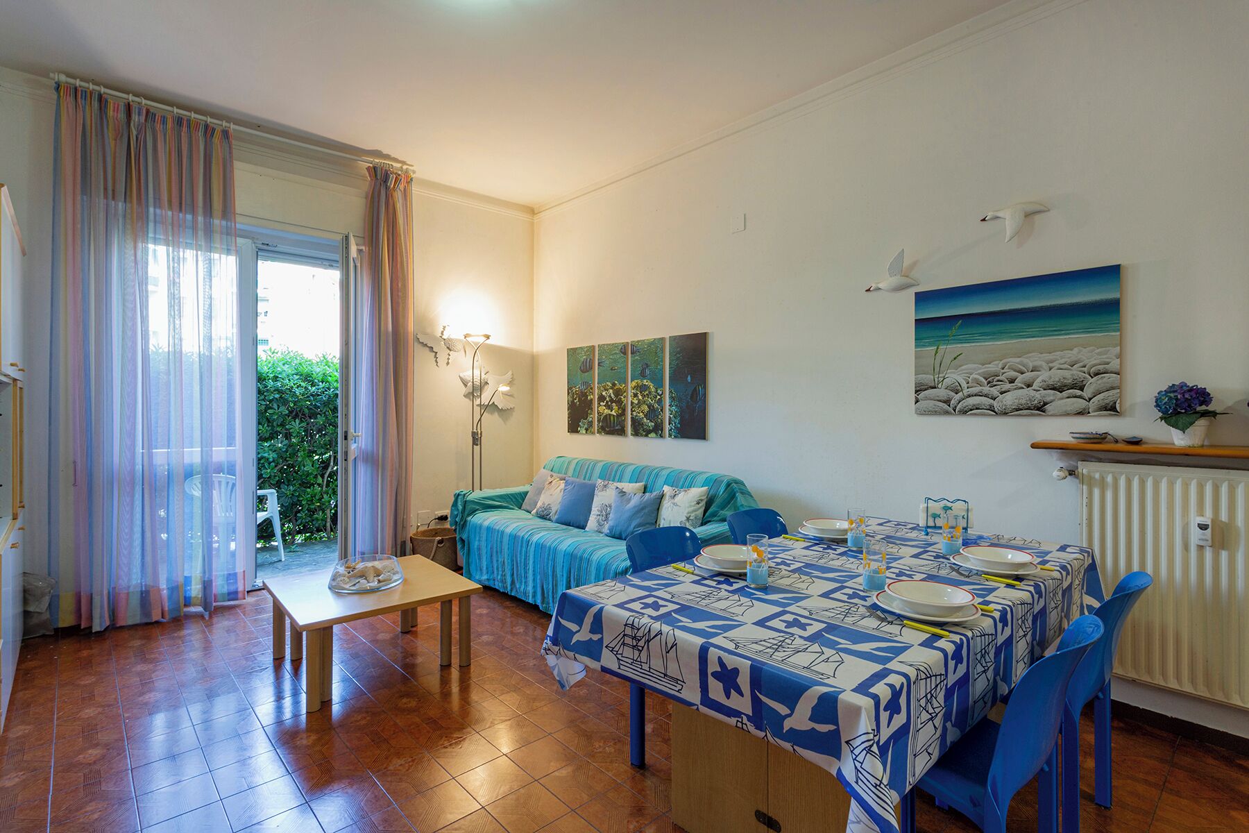 Une pièce comprenant une table à manger dressée pour quatre personnes, un canapé, un radiateur et un tableau représentant une scène de plage.