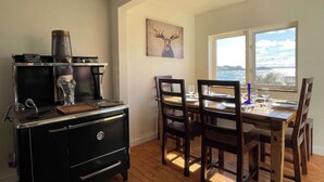 A dining area with a wooden table and chairs, a black stove, and a window with a view of the sea. - The Moose, Twillingate Island (Crow Head)