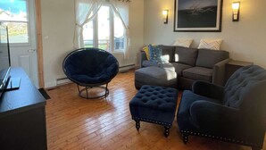 A living room with a dark blue chair, a grey sofa, and a matching ottoman. There is a window with white curtains, a framed picture on the wall, and a lamp on the side table. - The Moose, Twillingate Island (Crow Head)