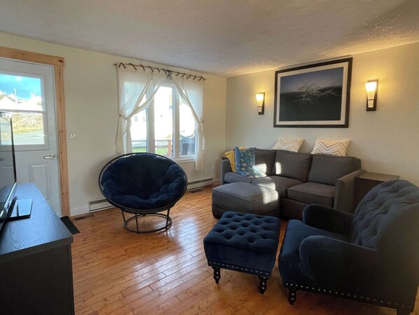 A living room with a dark blue chair, a grey sofa, and a matching ottoman. There is a window with white curtains, a framed picture on the wall, and a lamp on the side table. - The Moose, Twillingate Island (Crow Head)