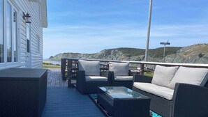 A deck with outdoor furniture, a glass table, and a view of the ocean and hills. - The Moose, Twillingate Island (Crow Head)