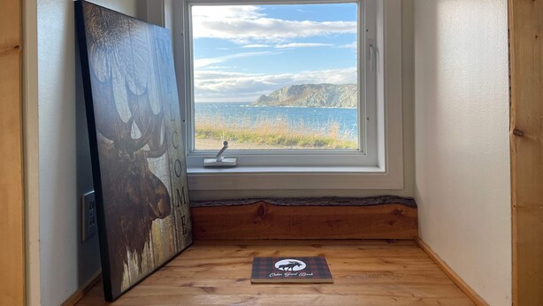 A corner room with a large window overlooking a coastal landscape, a rustic wooden floor, and a framed artwork on the wall. - The Moose, Twillingate Island (Crow Head)