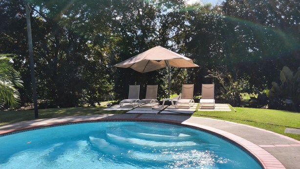 En swimmingpool med klart vand, omgivet af en murstensflisebelagt terrasse, og skyggefuld under en stor parasol.