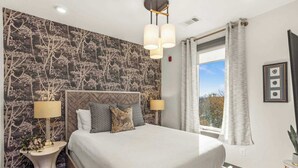 Cama bem arrumada com cabeceira, duas luminárias de apoio e uma janela com cortinas.