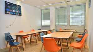 A conference room with multiple tables and chairs, a mounted TV on the wall, and large windows with blinds. - XENON HOSTELS UG (Dreieich)