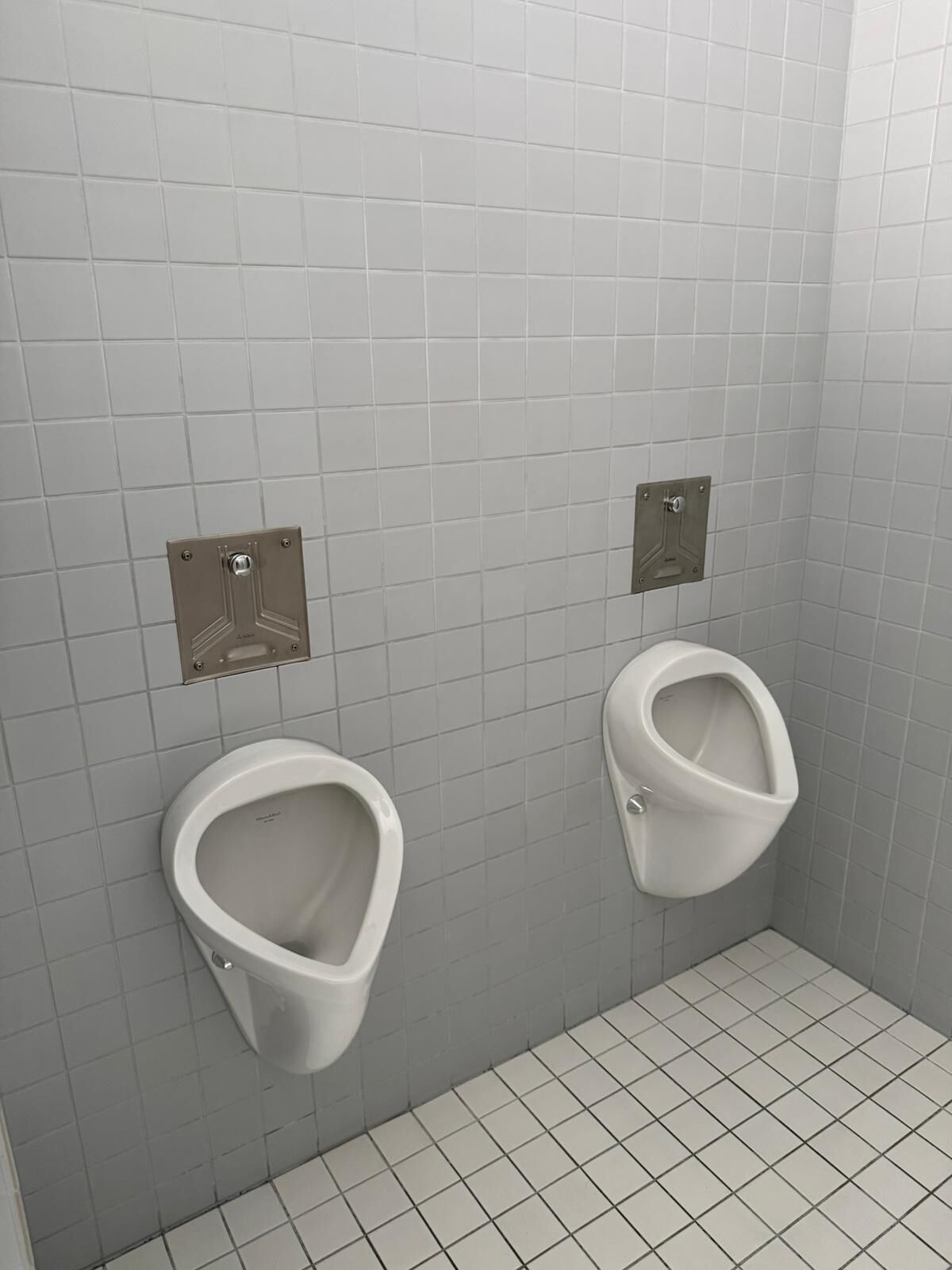 WC-tila, jossa on kaksi seinään kiinnitettyä, harmaalaatoitettujen seinien päällä olevaa pissaaria, ja kummankin pissaarin yläpuolella on metallinen huuhtelulevy. Lattia on laatoitettu.