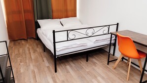 A single bed with a metal frame, a wooden desk with a chair, and a window with orange curtains. - XENON HOSTELS UG (Dreieich)
