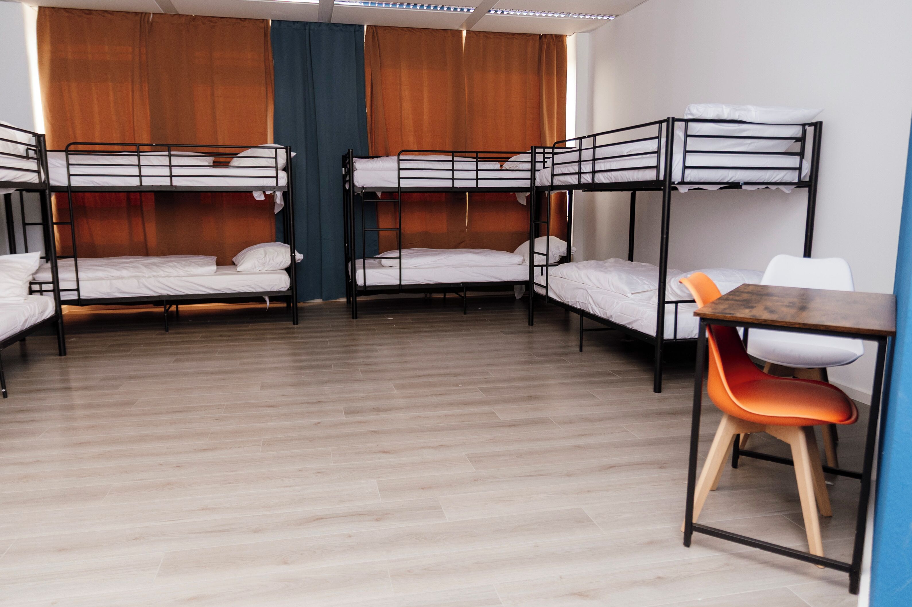 A dormitory room with bunk beds, a wooden floor, and a desk with a chair.
