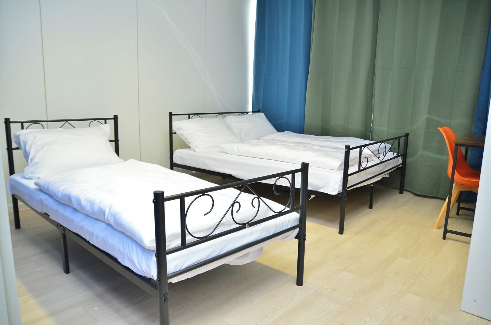 Zwei Einzelbetten mit Metallgestellen und weißer Bettwäsche in einem Zimmer mit Holzboden und einem grünen Vorhang.
