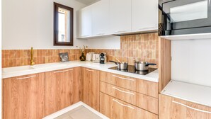 A modern kitchen with wooden cabinets, a white countertop, a built-in microwave, and a window with blinds. - La Maison de la Plage - Solea 3 Personnes (Mauguio)