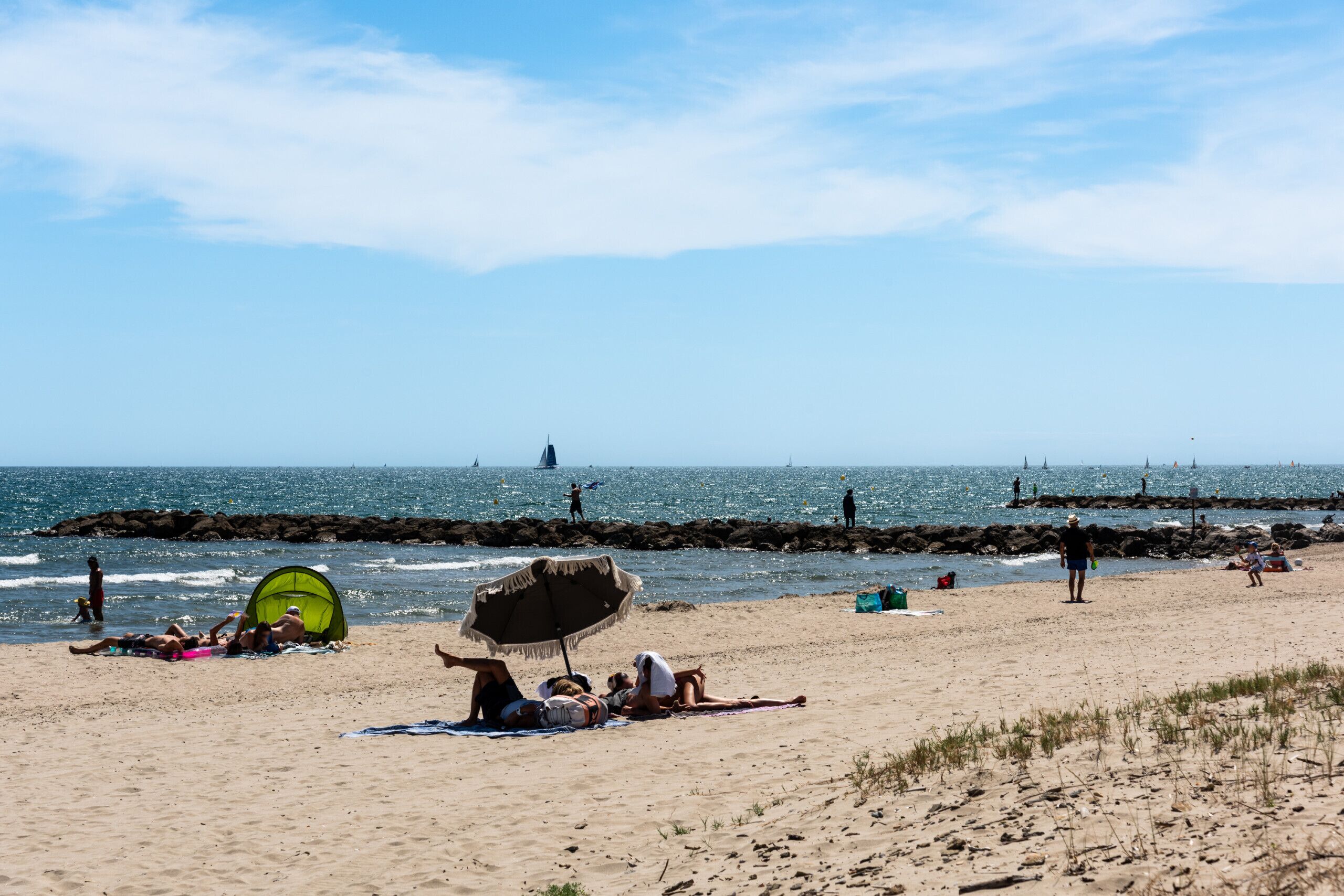 En strand med folk, der slapper af, en grøn parasol og en molle i baggrunden.