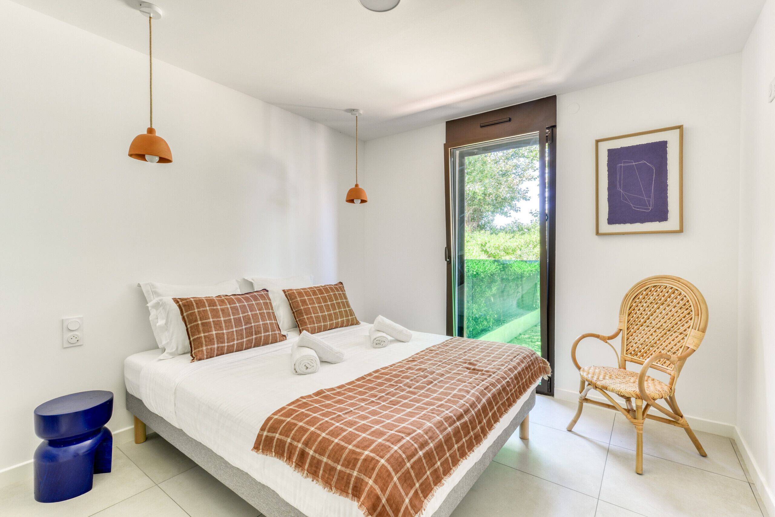 Camera da letto con un letto, due lampade a sospensione, una poltrona in vimini, un'opera d'arte incorniciata e vista panoramica attraverso una porta in vetro.