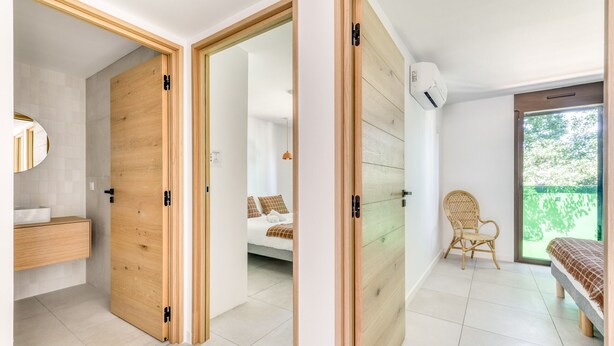 A modern bedroom with a wooden door, a bed, a chair, and a window with a view of greenery. - La Maison de la Plage - Alma 4 Personnes (Mauguio)