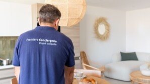 A person in a blue shirt with text on the back, standing in a modern living room with a dining table, chairs, and a sofa. - La Maison de la Plage - Alma 4 Personnes (Mauguio)