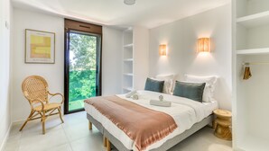A modern bedroom with a large bed, a wicker chair, and a view of greenery through a glass door. - La Maison de la Plage - Edena 4 Personnes (Mauguio)