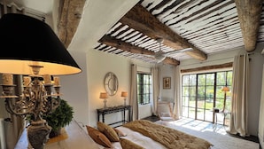 A bedroom with a large bed, a chandelier, a console table with lamps, a chair, and large windows. - Villa Hautvallon (Gordes)